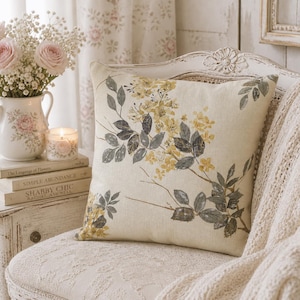 May include: A square cushion with a floral pattern of yellow flowers and grey leaves on a cream backdrop. The cushion rests on a white, ornate chair, with a cream-coloured throw. The scene has a vintage, cottagecore aesthetic.
