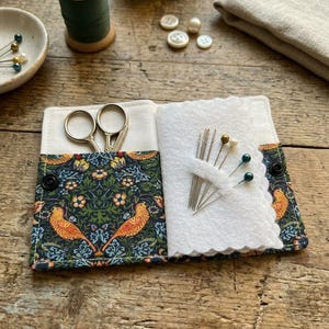 May include: A fabric needle case featuring a dark blue floral and bird pattern, containing silver scissors and a white felt page with several sewing needles. The case is secured with black snaps and includes a pocket for the scissors.
