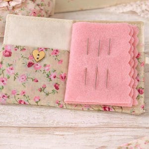 May include: Open floral fabric needle case with a pink felt interior holding several needles. The case has a heart-shaped wooden button closure and a ribbon tie. The fabric has a cream background with pink and red roses.
