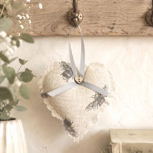 May include: A heart-shaped fabric ornament made of beige linen with a printed grey bee pattern. The ornament features a frayed edge, a grey ribbon tied in a bow at the top, and a small wooden heart-shaped button at the centre of the ribbon knot.