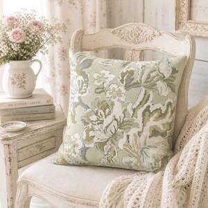 May include: A square cushion with a floral design in green, white, and blue sits on a white chair. The chair has a distressed finish. A vase of pink roses and books are on a side table.