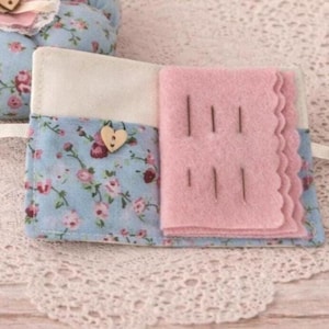 May include: A fabric needle book featuring a light blue floral print exterior with a small wooden heart button. The interior contains a cream-coloured pocket and a pink felt page with scalloped edges, holding several silver sewing needles.
