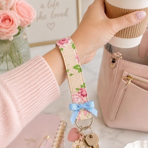 May include: A fabric wristlet keychain featuring a beige background with white polka dots and a pink floral pattern. The strap is accented with a small light blue ribbon bow and includes a silver metal ring holding two keys, a pink heart charm, and a flower charm.