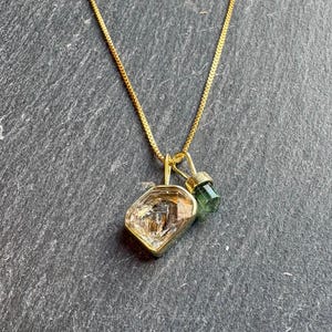 Peut inclure: Un collier à chaîne dorée avec deux pendentifs : une grande pierre transparente irrégulière et facettée avec des inclusions internes dans une monture dorée, et un plus petit pendentif en pierre verte translucide cylindrique avec un capuchon doré.