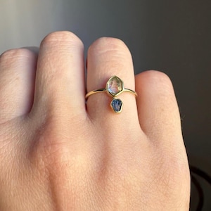 May include: A gold ring featuring two raw, uncut gemstones. The larger stone is a pale, translucent green with a hexagonal shape, while the smaller stone below it is a deep, saturated blue. Both stones are set in simple gold bezels on a thin band.