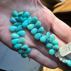 May include: A strand of faceted, teardrop-shaped turquoise beads in a vibrant cyan blue color. The beads are arranged in a cluster and attached to a light-colored rectangular tag with the handwritten number 190, ending in a matching cyan tassel.
