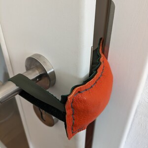 Könnte beinhalten: Ein Türstopper aus orangefarbenem Leder mit grünem Wildlederbesatz. Der Türstopper ist mit einem grünen Gummiband an einem Türgriff befestigt.
