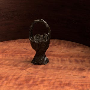 May include: A dark, textured basket with a handle, set on a wooden surface. The basket has an organic, almost tree-like appearance, with a rough, uneven texture and a dark, possibly bronze or charcoal color. The background is a dark, curved wooden panel.