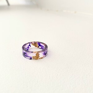 Op de afbeelding: Een ring van helder hars met paarse en gouden swirls. De ring is een statement piece met een uniek ontwerp.