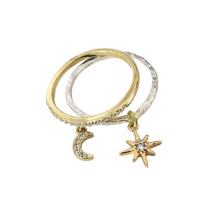 May include: Two rings, one gold and one silver, are intertwined. The gold ring has small clear stones and a crescent moon charm. The silver ring has a hammered texture and a star charm. Both charms have a small clear stone in the center.