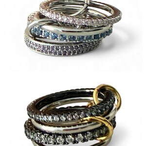 May include: Two stacks of rings. The top stack features silver rings with pink, blue, and purple gemstones. The bottom stack has silver rings with black and white gemstones, and gold accents. The rings are on a white background.