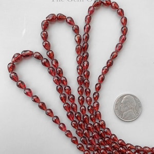 Könnte beinhalten: Eine Strähne tief roter Edelsteinperlen in Tropfenform. Die Perlen sind auf einem durchsichtigen Faden aufgefädelt und haben etwa die Größe einer deutschen Zehn-Cent-Münze.
