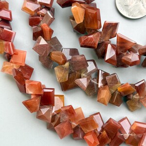 May include: Strands of faceted gemstone beads in shades of orange, red, and brown. The beads have a geometric, angular shape, and are strung together. The gemstones have a translucent quality, with visible internal patterns.