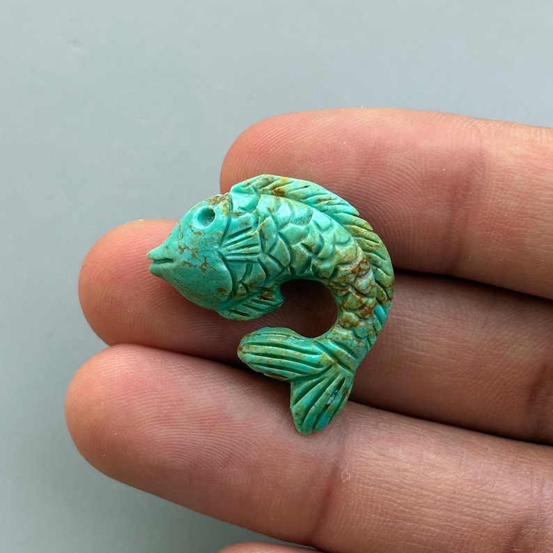 May include: A carved turquoise fish figurine with detailed scales and fins. The fish is a vibrant teal color with brown accents, and is held in a hand. The figurine is likely a decorative item or a piece of jewelry.