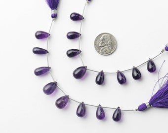 May include: A strand of purple amethyst gemstone beads in a teardrop shape. The beads are strung on a silver wire and are approximately 10mm in size. The beads are shown next to a US penny for size comparison. TheGemOrigin is printed on the image.
