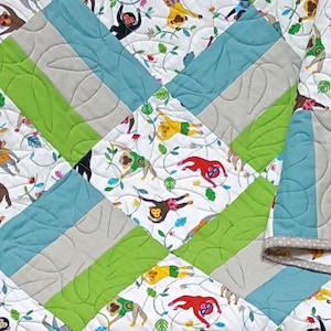 Op de afbeelding: Een witte, groene, blauwe en grijze babydeken met een apenpatroon. De deken is dubbelgevouwen en heeft een witte achtergrond met apen die aan wijnstokken hangen. De apen zijn in verschillende kleuren, waaronder rood, geel, blauw en groen.