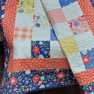 May include: A patchwork quilt with a white background and colorful squares. The squares feature floral patterns, gingham, and solid colors. The quilt is edged with a blue and white floral border.