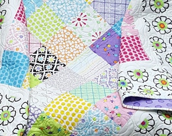 Op de afbeelding: Een kleurrijk patchwork-deken met een witte achtergrond en een rand van zwarte en groene bloemen. Het deken is gemaakt van vierkanten van verschillende stoffen, waaronder stippen, strepen en bloemenprints.