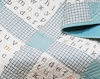 Op de afbeelding: Een wit dekbed met een blauw-wit geruit patroon. Het dekbed is gemaakt van vierkanten met geborduurde letters van het alfabet. De letters zijn in een willekeurig patroon gerangschikt. Het dekbed is dubbelgevouwen en ligt op een houten stoel.