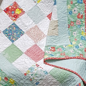 May include: A light blue and white quilt with a floral pattern. The quilt is made of patchwork squares and has a scalloped edge. The quilt is folded and displayed on a wooden chair.