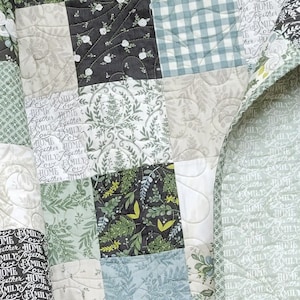 May include: A green, white and black patchwork quilt with a floral pattern and the words "Love Family Home Together" repeated throughout. The quilt is folded in half and the edges are sewn with a white trim.