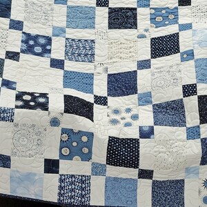 May include: A blue and white patchwork quilt with a variety of patterns, including sunbursts, dots, and swirls. The quilt is made of squares in shades of blue and white, creating a visually appealing design. The quilt is likely made of cotton.