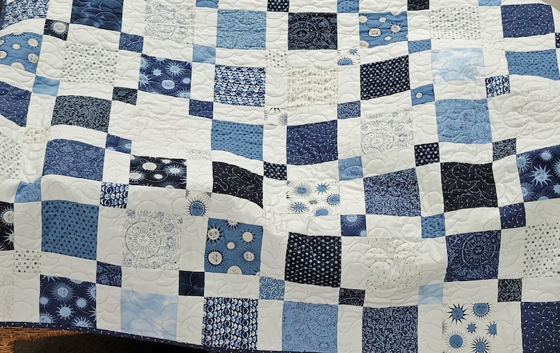 May include: A blue and white patchwork quilt with a variety of patterns, including sunbursts, dots, and swirls. The quilt is made of squares in shades of blue and white, creating a visually appealing design. The quilt is likely made of cotton.