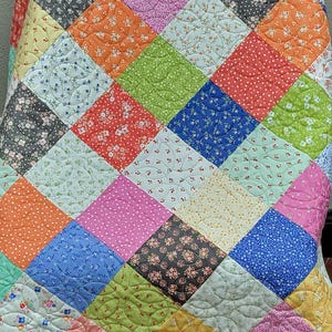 Può includere: Una colorata trapunta patchwork con una varietà di quadrati a motivi nei toni del blu, rosa, arancione, verde e giallo. La trapunta ha un design trapuntato e un bordo verde. La trapunta è probabilmente fatta a mano.