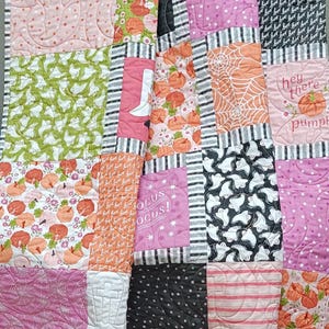 Op de afbeelding: Een patchworkquilt met een Halloween-thema. De quilt heeft verschillende patronen, waaronder pompoenen, geesten, spinnenwebben en de tekst "hey there pumpkin". Kleuren zijn roze, oranje, groen, zwart en wit.