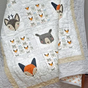 May include: A handmade baby quilt featuring a repeating pattern of woodland animals, including foxes, owls, and bears. The quilt is primarily white with gray accents and a beige border. The animals are appliqued onto the quilt.