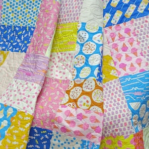 Könnte beinhalten: Ein farbenfroher Patchwork-Quilt mit verschiedenen Mustern, darunter Eiswaffeln, Süßigkeiten und Tupfen. Der Quilt zeigt eine Mischung aus rosa, blauen, gelben und orangefarbenen Quadraten, die ein verspieltes Design ergeben. Der Quilt ist mit einem Wellenmuster gesteppt.