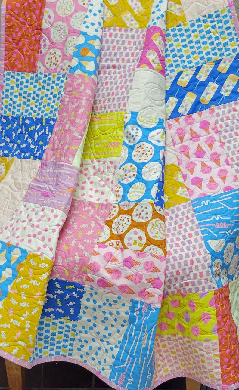 Pu&ograve; includere: Una colorata trapunta patchwork con vari motivi, tra cui coni gelato, caramelle e pois. La trapunta presenta un mix di quadrati rosa, blu, gialli e arancioni, creando un design giocoso e allegro. La trapunta &egrave; trapuntata con un motivo ondulato.