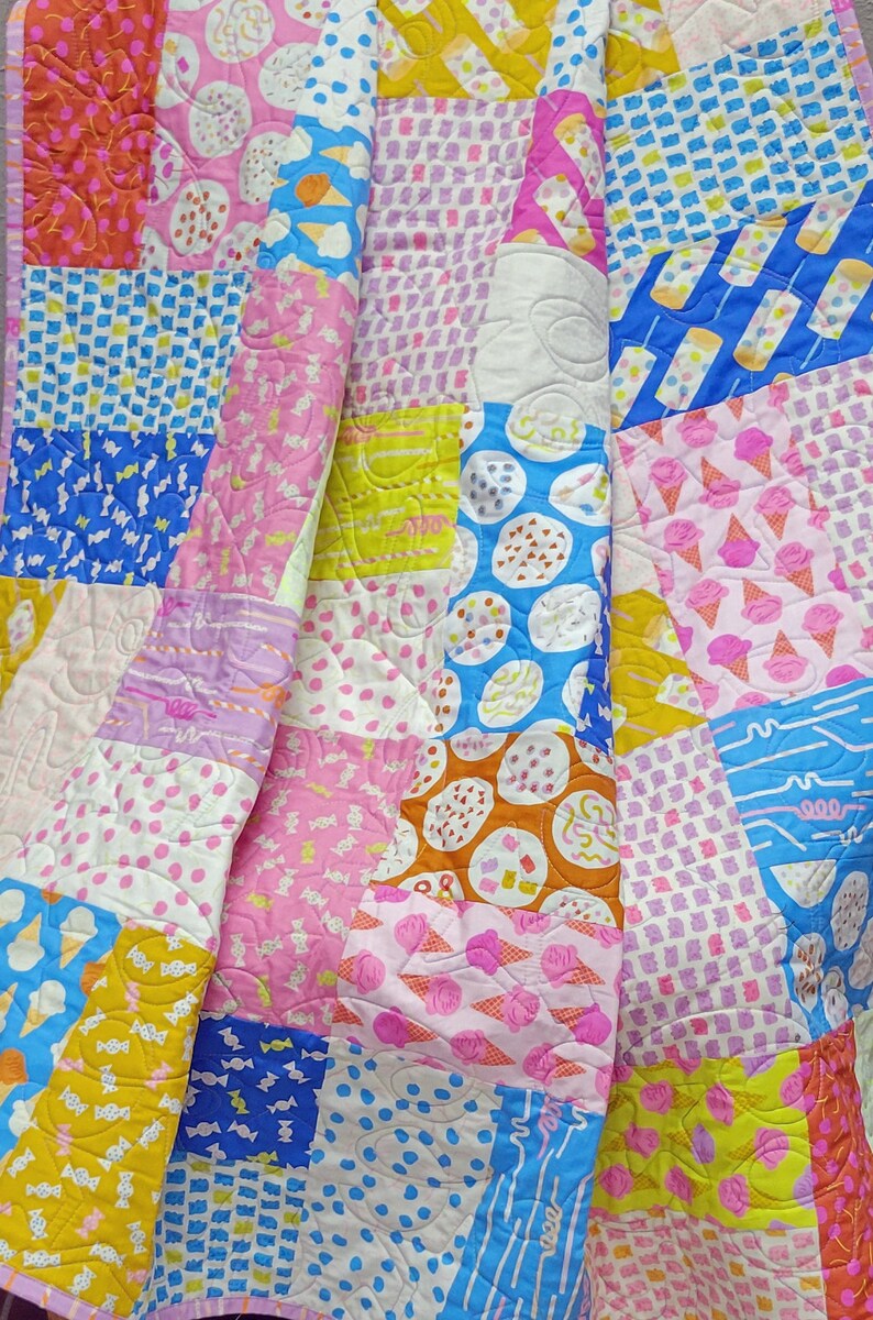 Pu&ograve; includere: Una colorata trapunta patchwork con una variet&agrave; di motivi, tra cui coni gelato, caramelle e pois. La trapunta presenta un mix di tessuti rosa, blu, gialli e arancioni, creando un design allegro e giocoso.