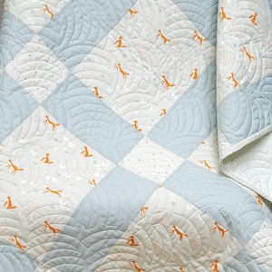 May include: A white and blue baby quilt with a checkered pattern and a fox print. The quilt is quilted with a wavy pattern.