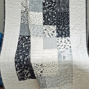 Puede incluir: Una colcha de retazos en blanco y negro con una variedad de patrones, incluyendo diseños florales, geométricos y abstractos. La colcha está doblada y se muestra en una silla de madera.