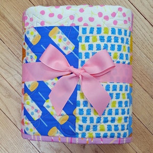 Pu&ograve; includere: Una trapunta piegata con un nastro rosa legato a fiocco. La trapunta presenta motivi colorati di ghiaccioli, orsetti gommosi e ciliegie. La trapunta misura circa 76 cm di lunghezza e 63 cm di larghezza.