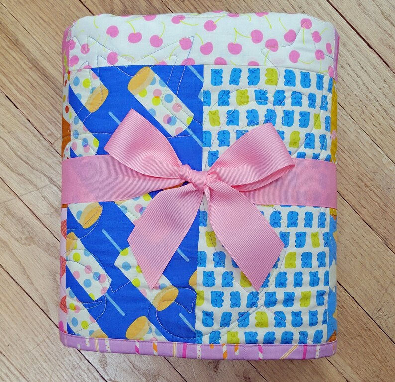 Pu&ograve; includere: Una trapunta piegata con un nastro rosa legato a fiocco. La trapunta presenta motivi colorati di ghiaccioli, orsetti gommosi e ciliegie. La trapunta misura circa 76 cm di lunghezza e 63 cm di larghezza.