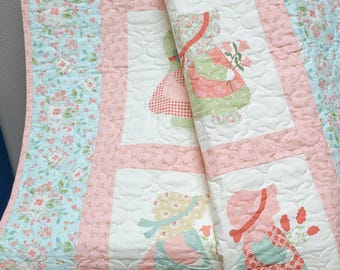 Puede incluir: Un edredón de colores pastel con un estampado floral y figuras aplicadas de niños con gorros y vestidos. El edredón presenta un borde floral azul claro, detalles en rosa y cuadrados blancos con las figuras aplicadas. El edredón está acolchado con un patrón ondulado.