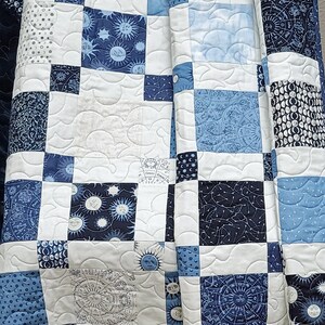 May include: A handmade quilt featuring a patchwork design in shades of blue and white. The squares display celestial patterns, including suns, moons, stars, and zodiac symbols. The quilt is machine-stitched with a wavy pattern.