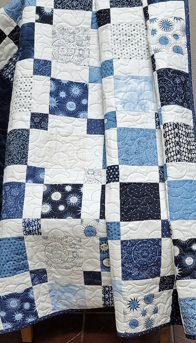 May include: A handmade quilt featuring a patchwork design in shades of blue and white. The squares display celestial patterns, including suns, moons, stars, and zodiac symbols. The quilt is machine-stitched with a wavy pattern.
