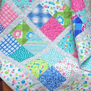 Könnte beinhalten: Eine Patchwork-Decke mit einem Rautenmuster in Rosa-, Blau-, Grün- und Weißtönen. Die Decke besteht aus verschiedenen Stoffen mit unterschiedlichen Mustern, darunter Punkte, Streifen und Blumenmuster.