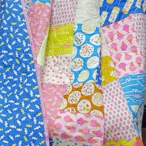 Pu&ograve; includere: Una colorata trapunta patchwork con vari motivi, tra cui caramelle, coni gelato e pois. La trapunta presenta un retro blu a fantasia caramelle e un bordo rosa. Il design incorpora rosa, blu, giallo e bianco.