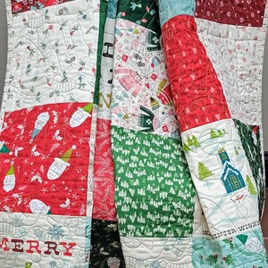 以下が含まれることがあります： さまざまなお祝いのパターンが施されたクリスマスをテーマにしたキルト。キルトは、雪だるま、セーター、クリスマスツリーの画像が描かれた赤、緑、白のパネルが特徴です。「MERRY & BRIGHT」の文字も表示されています。