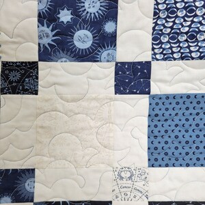 May include: A close-up of a quilted fabric with a celestial theme. The quilt features squares of navy blue and off-white fabric. The navy squares have sun, moon, and star patterns. The off-white squares have a cloud-like pattern and astrological designs.