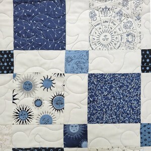 May include: A patchwork quilt featuring squares of white, navy blue, and light blue fabric. The quilt has a celestial theme, with suns, stars, and zodiac symbols. The squares are stitched together with white thread in a wavy pattern.