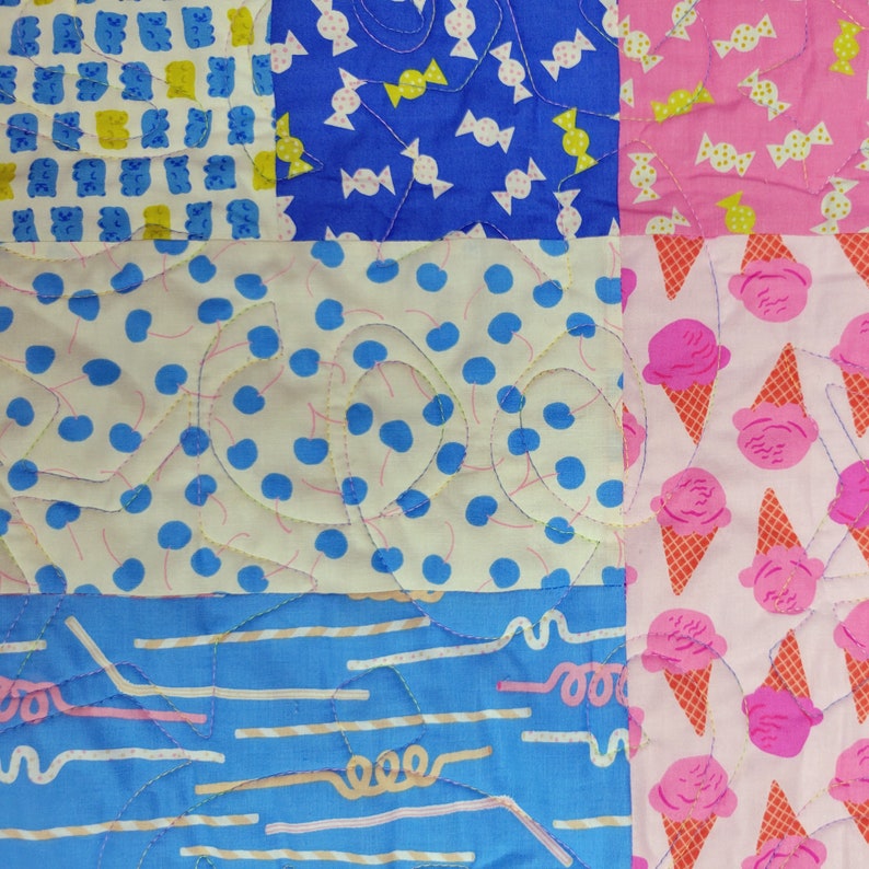Pu&ograve; includere: Una colorata trapunta patchwork con vari motivi. La trapunta presenta quadrati con orsetti gommosi blu, caramelle bianche su blu, caramelle rosa, pois blu, coni gelato rosa e linee e vortici blu. La trapunta &egrave; trapuntata a macchina con filo giallo.