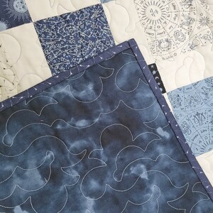 May include: A close-up of a handmade quilt with celestial and astrological patterns. The quilt features squares of white, navy, and light blue fabric. The backing is a dark blue fabric with a cloud-like pattern, and the edges are bound with a navy blue trim.