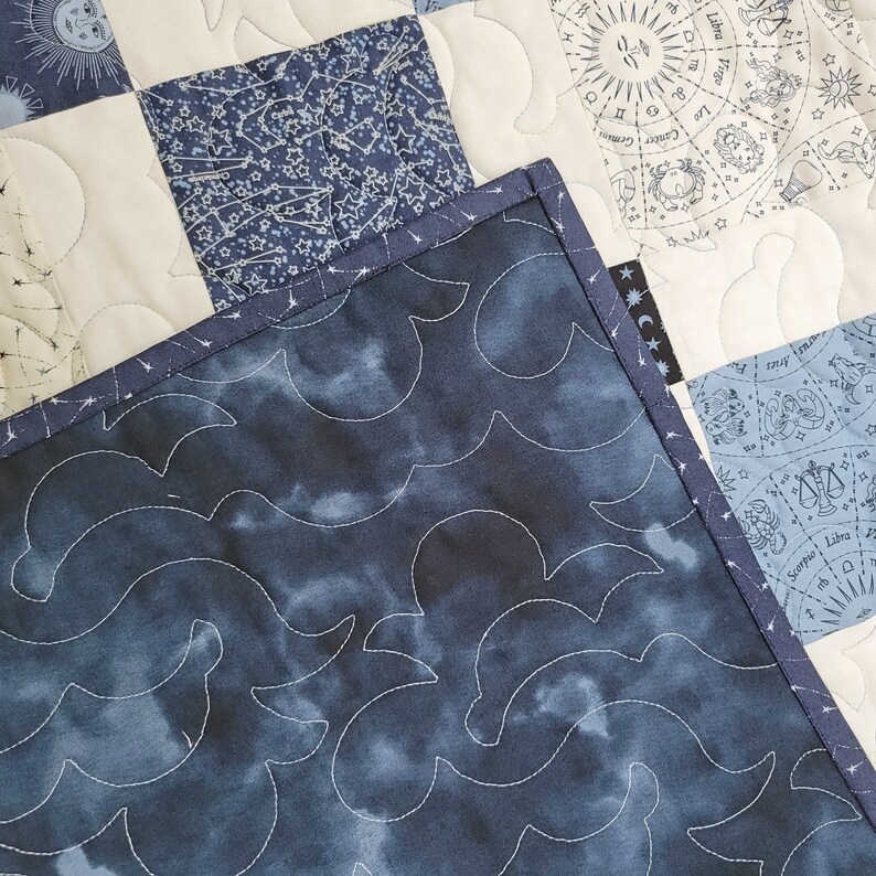 May include: A close-up of a handmade quilt with celestial and astrological patterns. The quilt features squares of white, navy, and light blue fabric. The backing is a dark blue fabric with a cloud-like pattern, and the edges are bound with a navy blue trim.