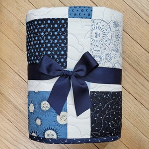 May include: A quilted blanket with a navy blue bow. The blanket is made of squares with celestial patterns in shades of blue and white. The patterns include moons, stars, and zodiac symbols. The blanket is tied with a wide navy blue ribbon.