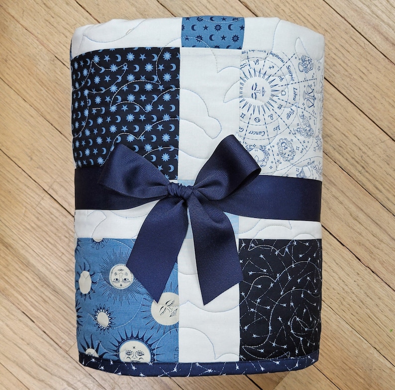 May include: A quilted blanket with a navy blue bow. The blanket is made of squares with celestial patterns in shades of blue and white. The patterns include moons, stars, and zodiac symbols. The blanket is tied with a wide navy blue ribbon.
